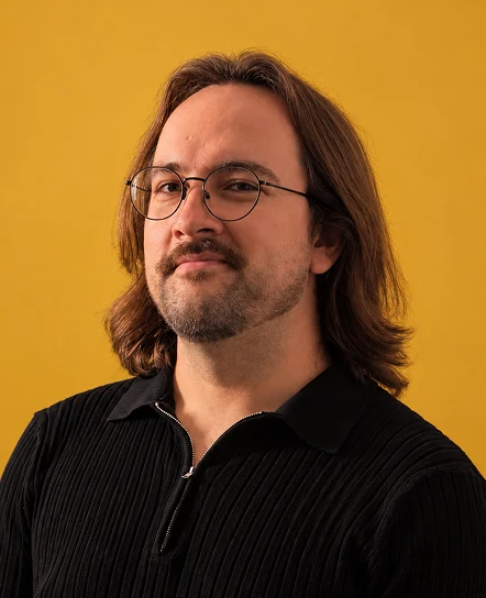 James, in glasses and a black shirt, against a mustard wall