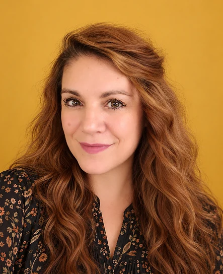 Megan, smiling, with long auburn hair, against a mustard wall